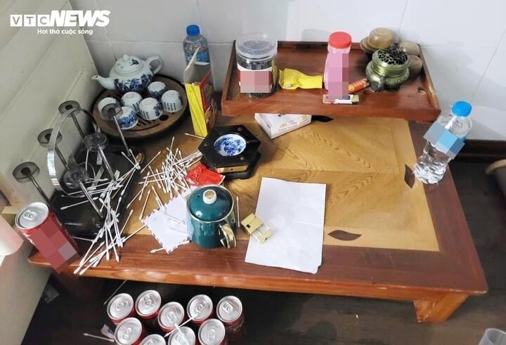 Góc phòng, chiếc tủ nhựa bung ngăn, chứa đầy vỏ thuốc, thực phẩm chức năng, đồ cá nhân lộn xộn. Bên cạnh là bàn gỗ bày biện đủ loại vật dụng phục vụ ăn chơi như ấm nước, hộp đồ mỹ phẩm, bình trà, lọ nhựa, khay đựng trà và thuốc, vỏ hộp thực phẩm chức năng.